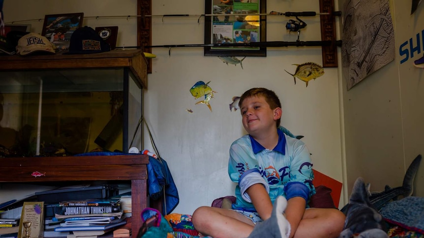 A young boy sits cross-legged on his bed smiling with fish stickers on the wall and a fish tank next to him