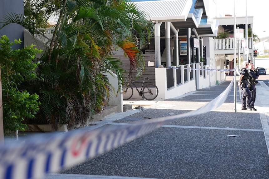 Police tape lines a street where two police officers stand in the morning sun. 