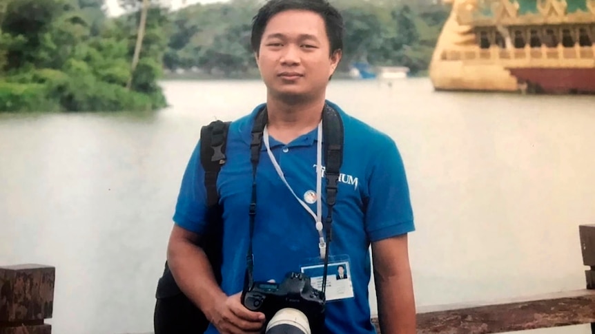 Thein Zaw wearing a blue polo shirt and holding a camera standing in front of a body of water