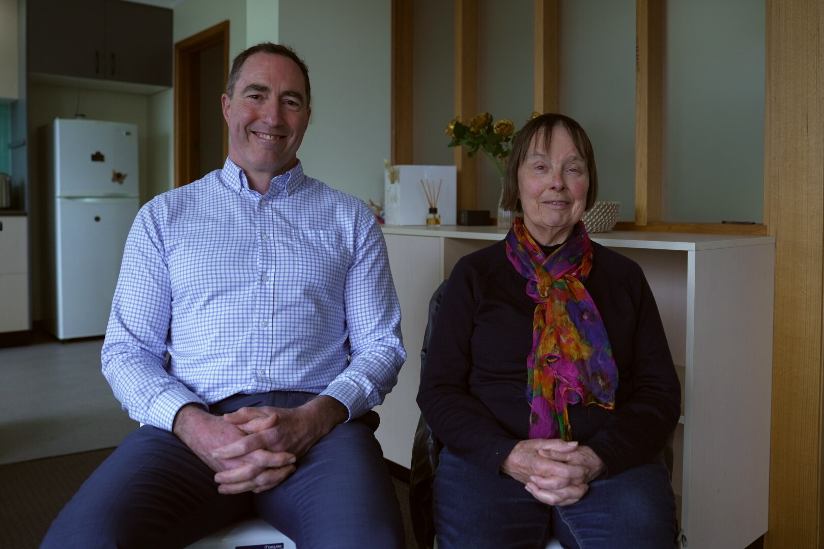 Un hombre con camisa y pantalones de negocios y una mujer con un corte bob corto y una bufanda de colores se sientan uno al lado del otro y sonríen.
