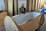 A man with a very long beard and no hair leans over an open coffin like structure 