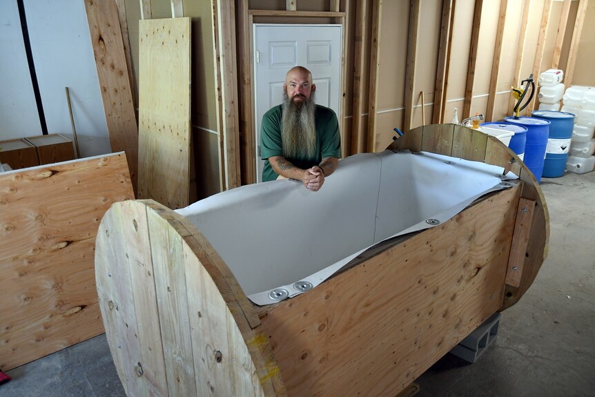 A man with a very long beard and no hair leans over an open coffin like structure 