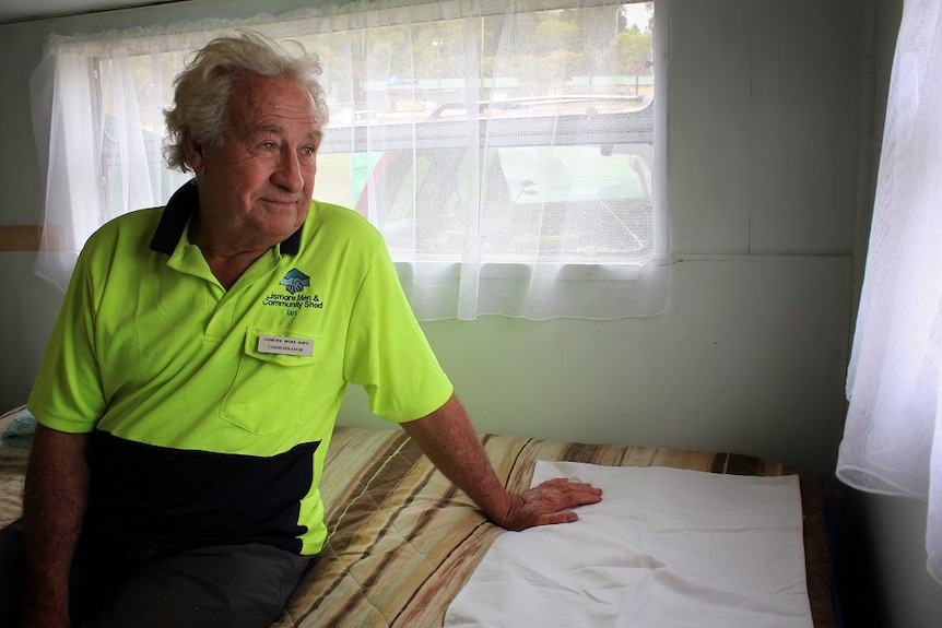 An older man sits on a bed in a caravan, looking out the window.