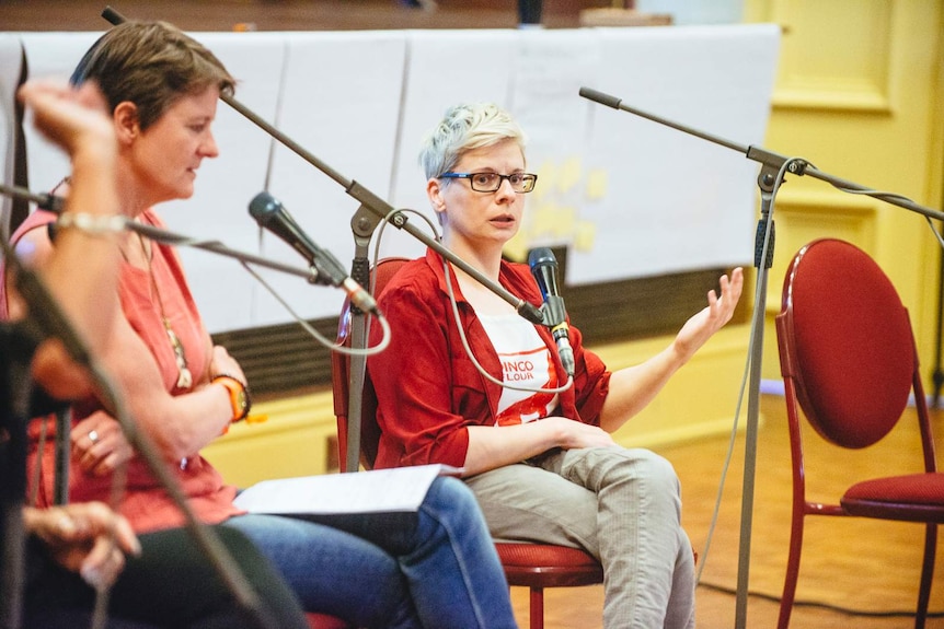 Woman with short platinum blonde hair and glasses in red hoodie, sitting in circle of others, microphones in frame.