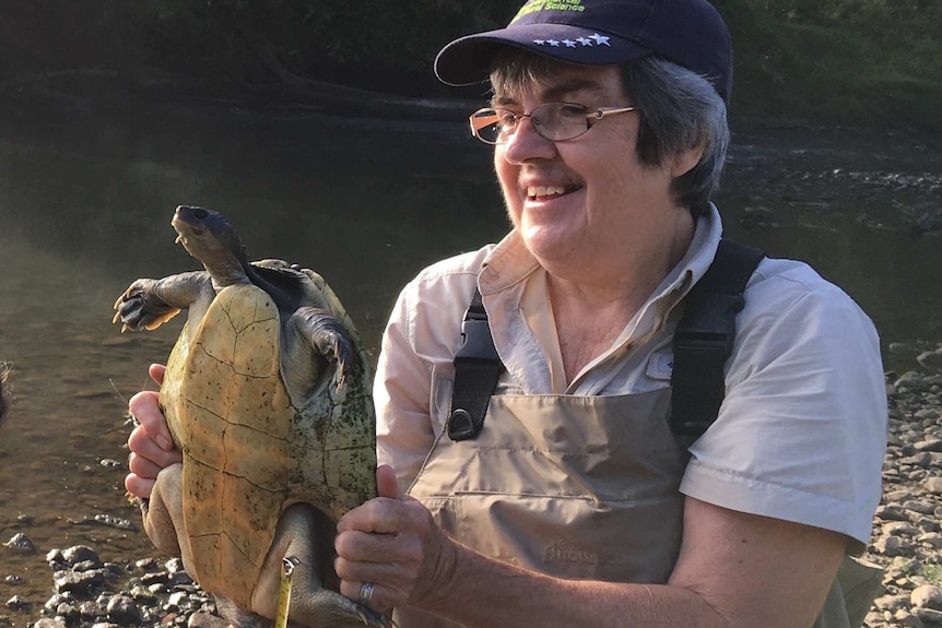 A smiling, middle-aged woman sits on a riverbank, holding a turtle.