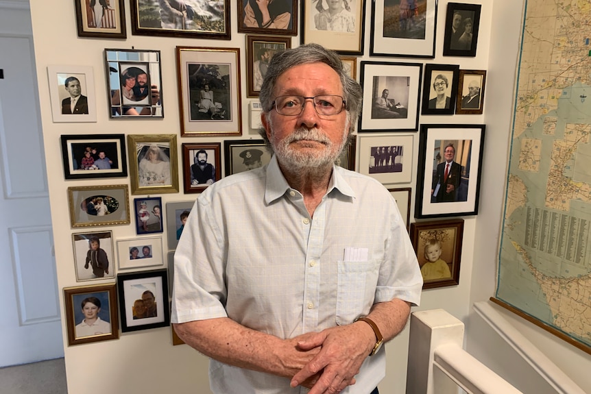 A man with grey hair and glasses stands in front of a wall of old photos. He's not smiling.