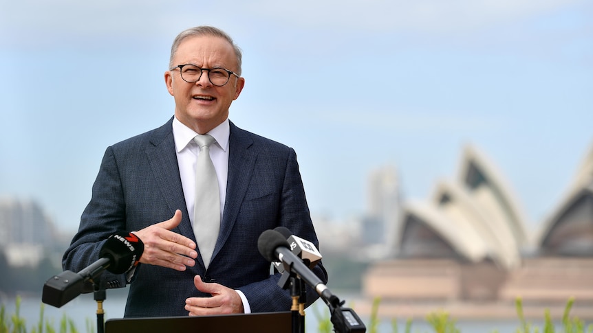 A middle-aged man in a suit with glasses speaks behind a microphone-laden lectern in front of the Sydney Opera House.