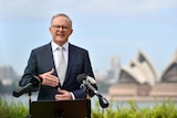A middle-aged man in a suit with glasses speaks behind a microphone-laden lectern in front of the Sydney Opera House.