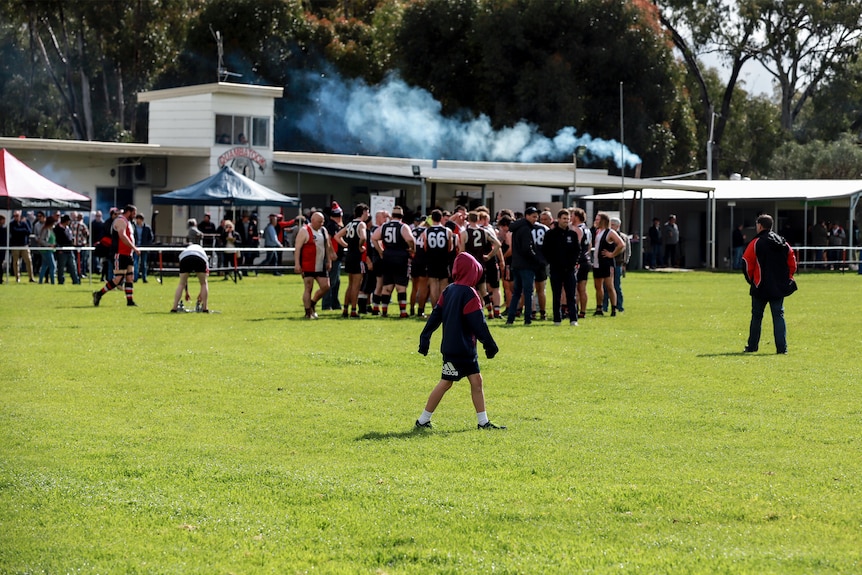 A child wearing shorts and a hooded jumper walks across a football oval with a pack of older football players in distance