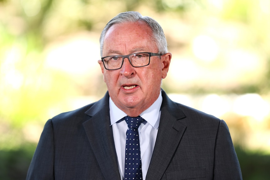 A man wearing glasses and a suit faces forward speaking