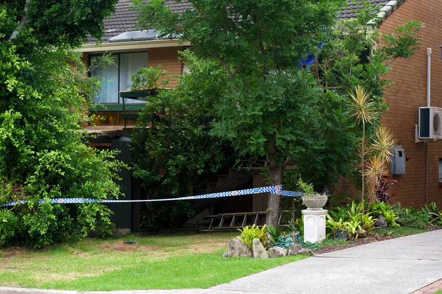 A brick house surrounded by lush green trees, with blue and white police tape strung up in the yard.