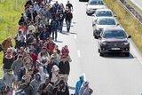 Elevated view of large group of asylum seekers walking beside a highway towards the photographer, with cars passing by.