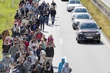 Elevated view of large group of asylum seekers walking beside a highway towards the photographer, with cars passing by.