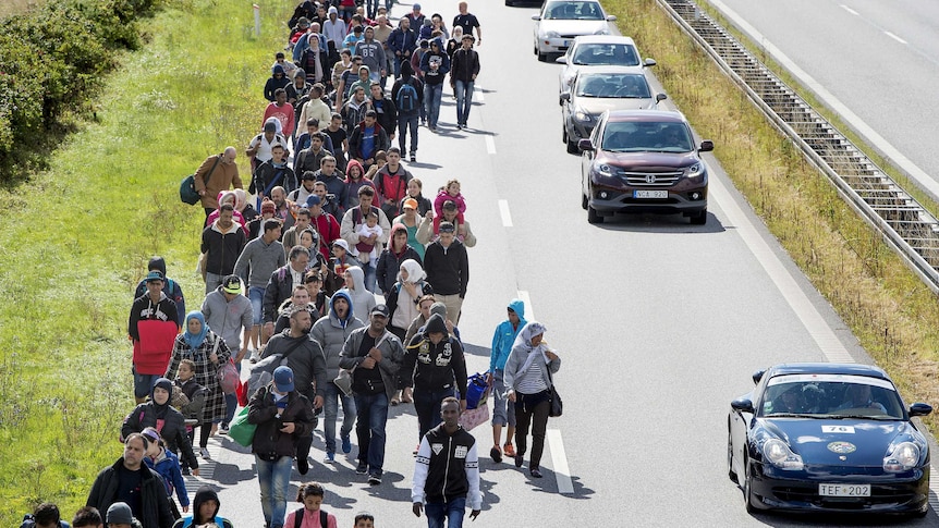 Elevated view of large group of asylum seekers walking beside a highway towards the photographer, with cars passing by.