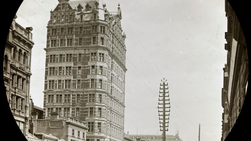 A black and white image of a 12-storey brick building with a gabled roof. People and tram in street in front.