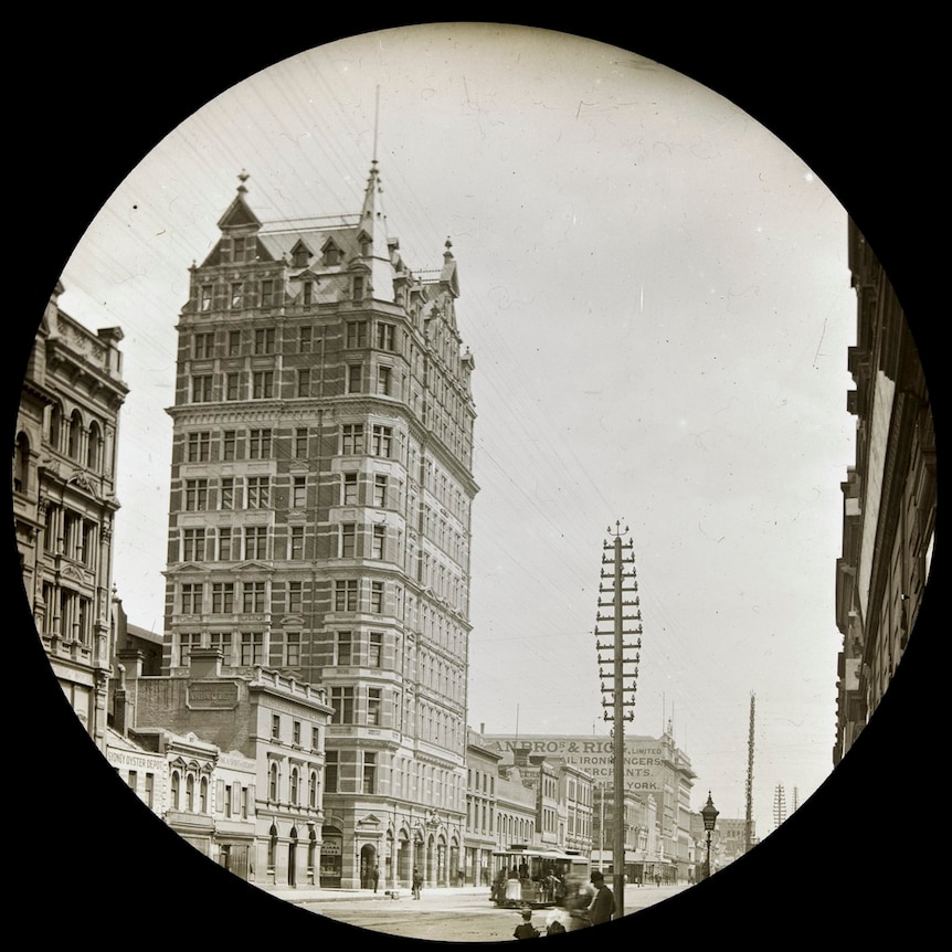 A black and white image of a 12-storey brick building with a gabled roof. People and tram in street in front.