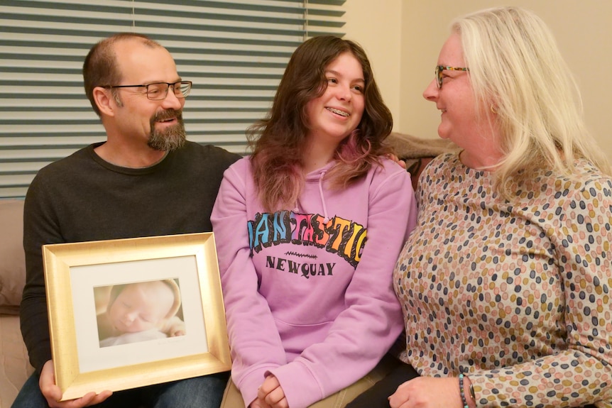 A mother and father sit on either side of a teenage girl smiling, while holding a baby photo