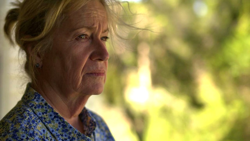 A close up of a woman staring ahead with a neutral expression, standing on the deck of a house, trees can be seen behind her.