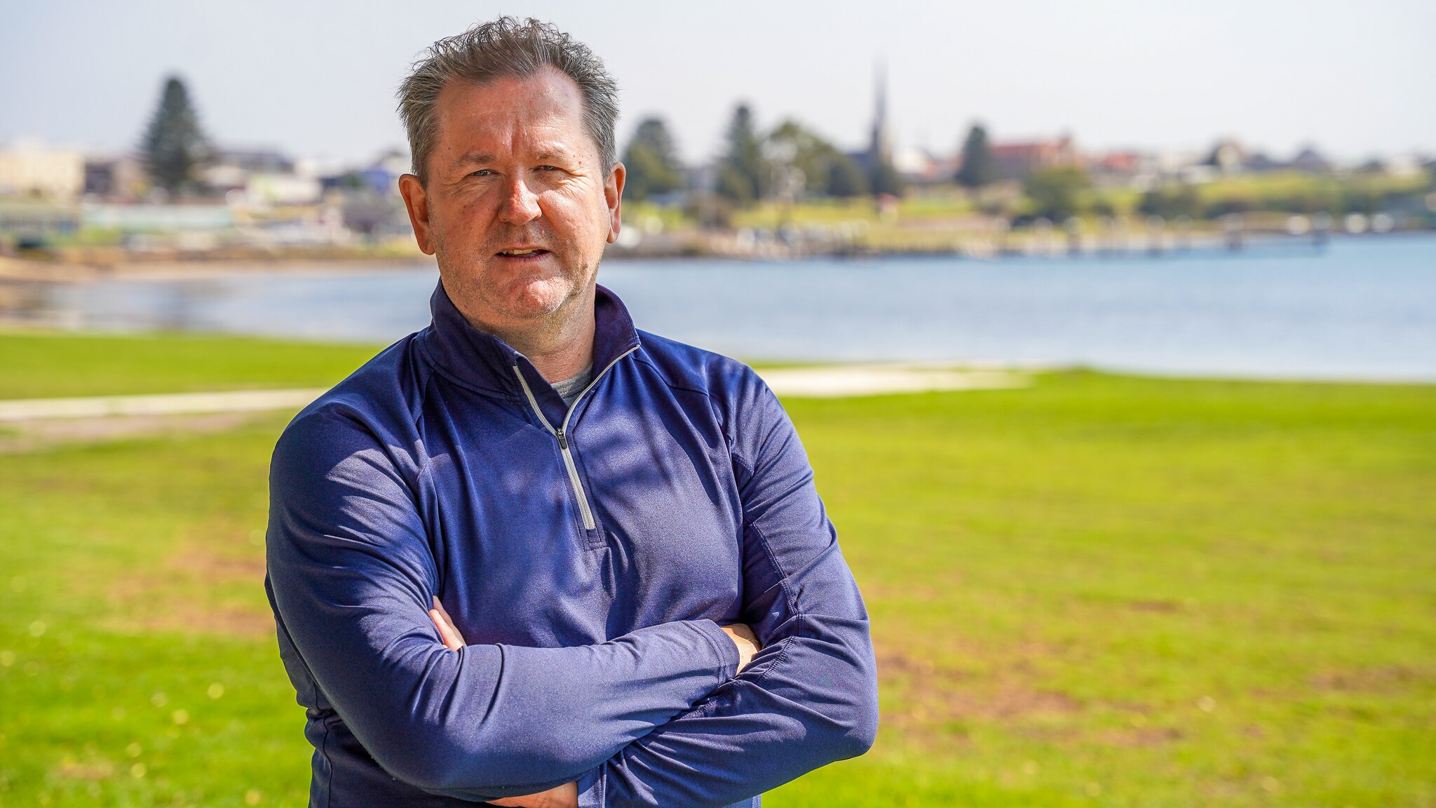 A man wearing a blue zipper sweater stands on a large patch of grass with his arms crossed.