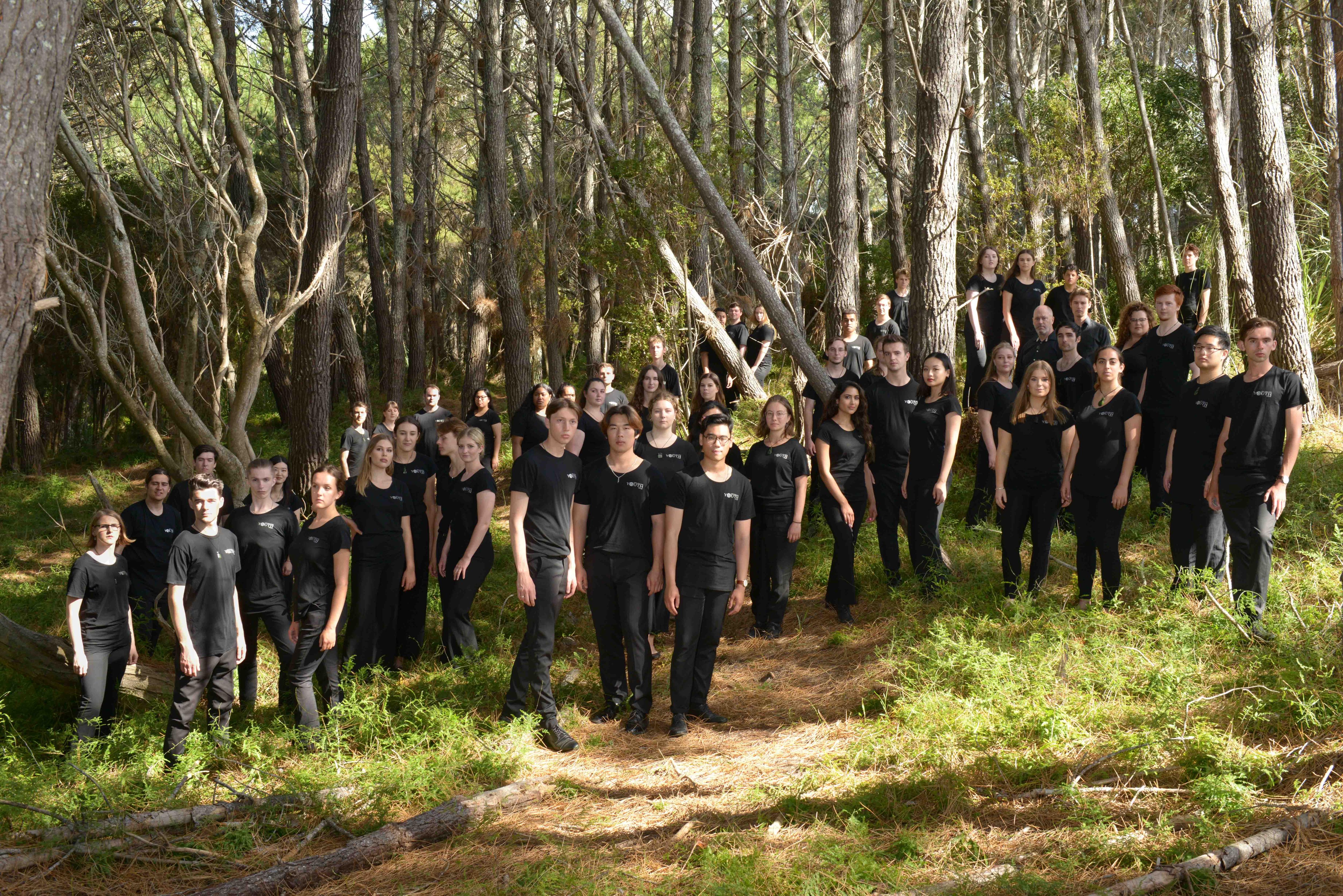 A group of about fourty people 18-25 years old standing amongst trees and a path. They are all wearing black.
