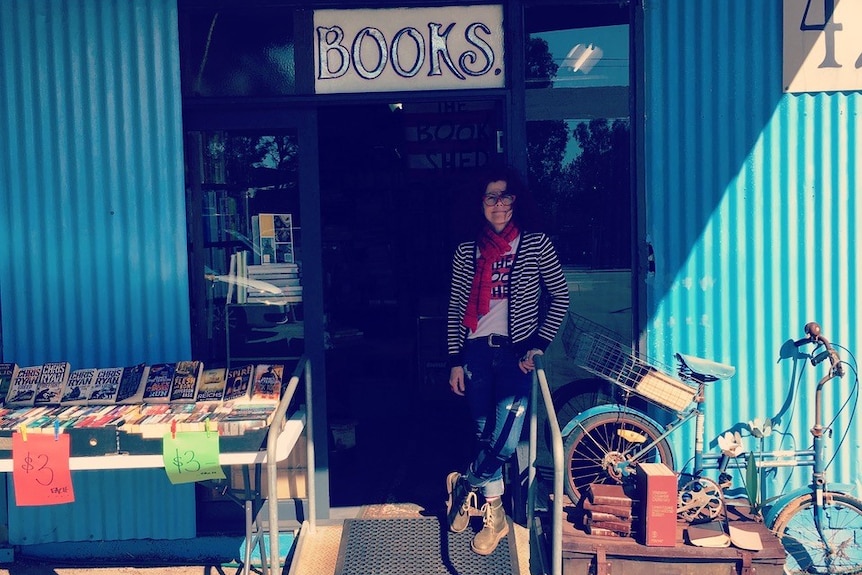 Kaz morgan stands outside a door that says 'Books' attached to a blue shed. There are books on the left and a bike on the right.