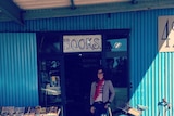Kaz morgan stands outside a door that says 'Books' attached to a blue shed. There are books on the left and a bike on the right.