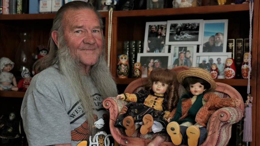 Man with long grey hair, sideburns with tattoos holding two dolls on little wooden couch in front of bookshelf of dolls photos