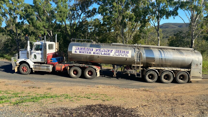 A truck towing a water tanker on a bitumen road with trees behind it