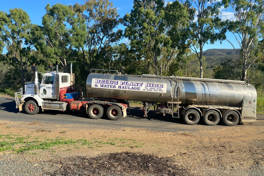 A truck towing a water tanker on a bitumen road with trees behind it