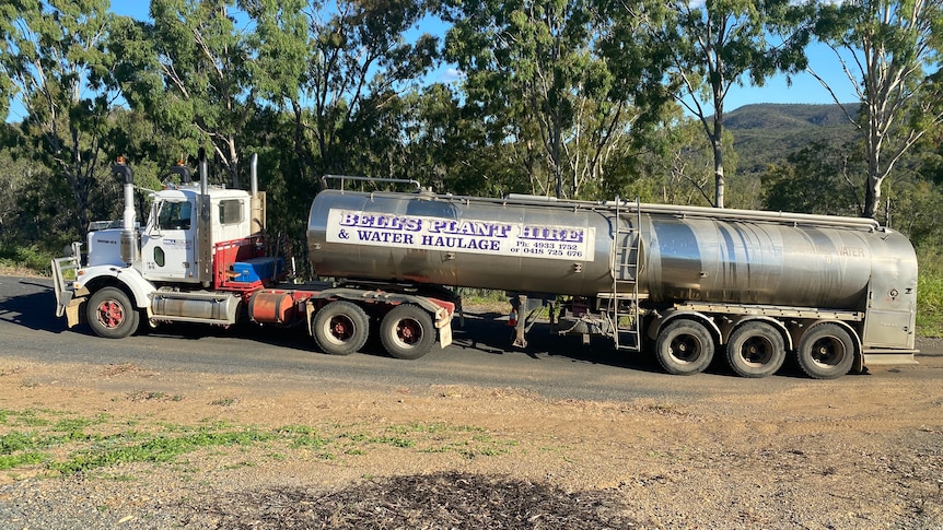 A truck towing a water tanker on a bitumen road with trees behind it