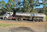 A heavy haulage water tanker on a narrow bitumen road with trees behind it and dusty roadside in foreground