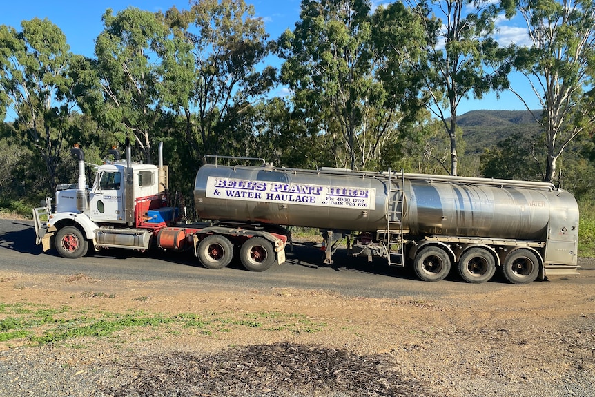 A truck towing a water tanker on a bitumen road with trees behind it