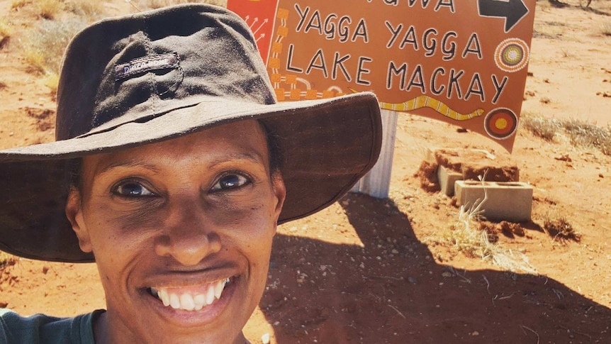woman in wide brimmed hat in front of sign to communities painted on an old car bonnet.