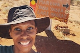 woman in wide brimmed hat in front of sign to communities painted on an old car bonnet.