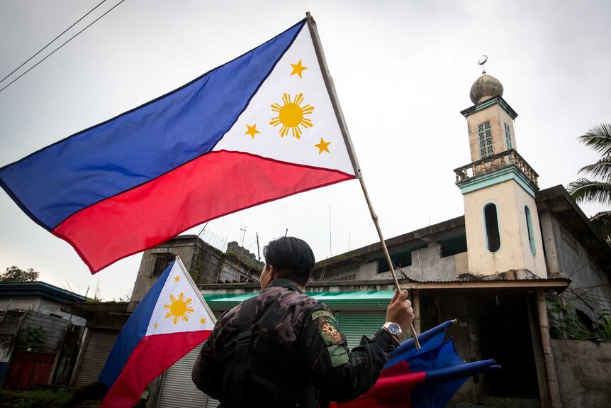 A soldier, with his back to the camera, holds a Filipino flag aloft, standing in front of a mosque.