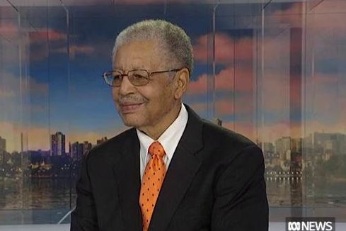 A smiling, bespectacled older man in formal attire sits in a TV studio