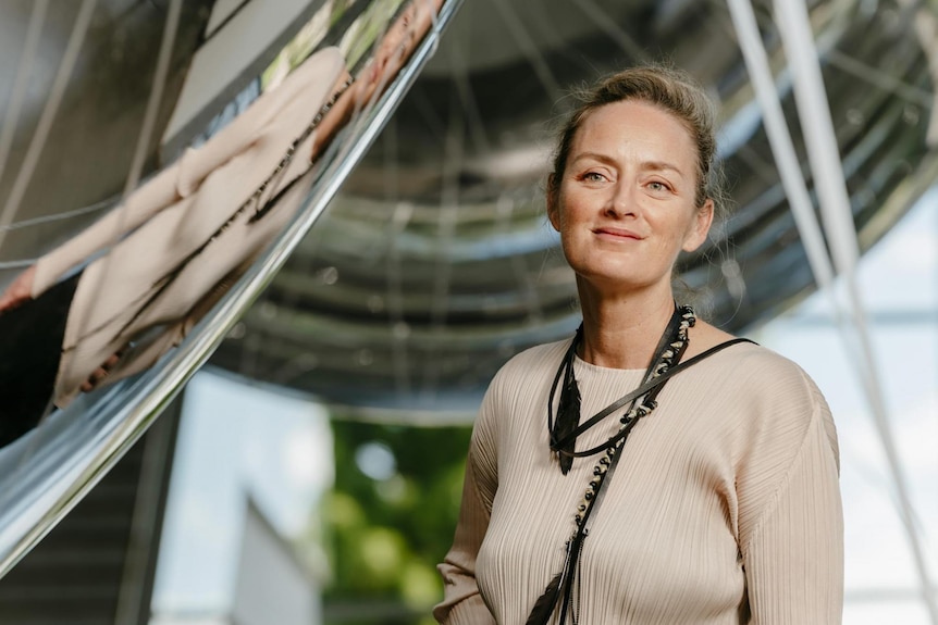 A middle-aged blonde woman stands in a light-filled atrium, smiling slightly. She is in front of two large silver spheres.