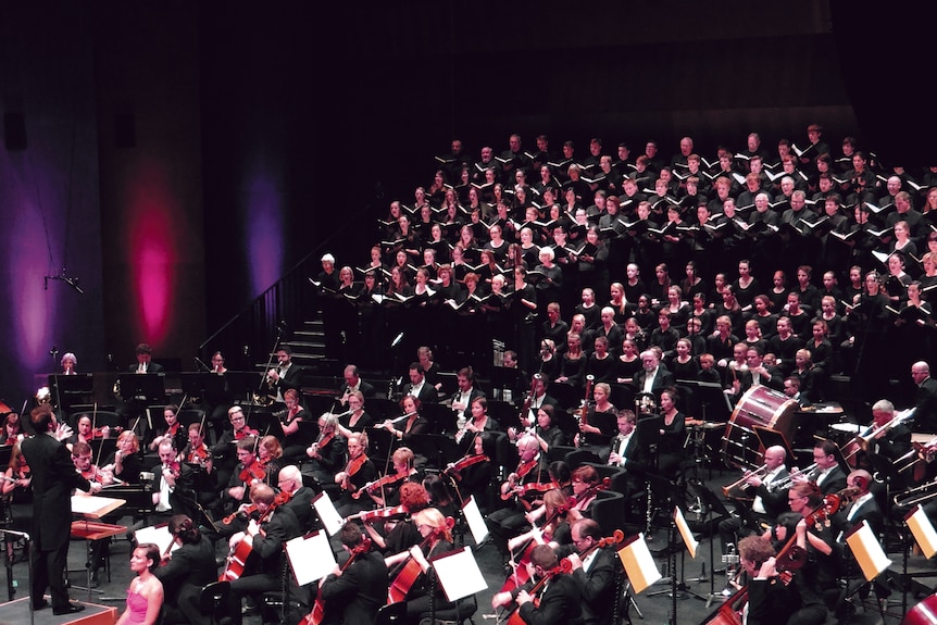 A choir and orchestra perform in front of a conductor on a stage