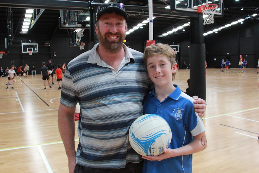 a father hugs his young son who is holding on a netball as they stand on a netball court