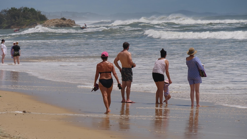 Multiple people walk along the beach. In the background a person rides a jetski in the surf. 