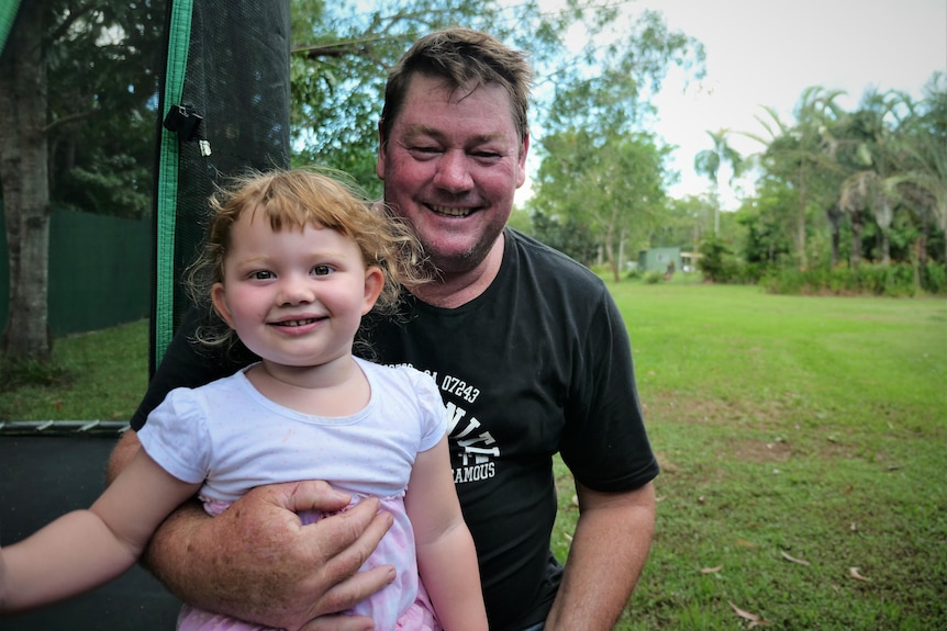 Father sitting on side of a garden trampoline with daughter on lap.