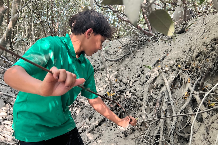 A boy in a green shirt poking a wire into a sandy creek bed.