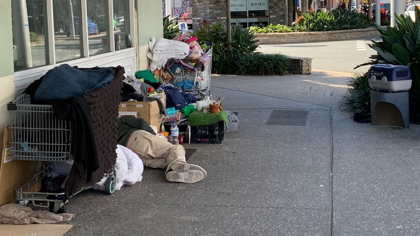 A man lies on the footpath surrounded by some shopping trolleys full of his possessions