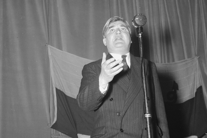 Black and white photo of a man giving a speech in front of a flag.