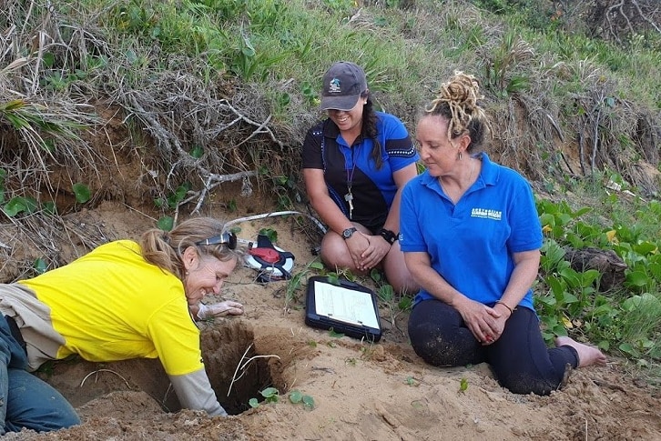 A group of women sit on the sand and watch another one digging a big hole in the ground