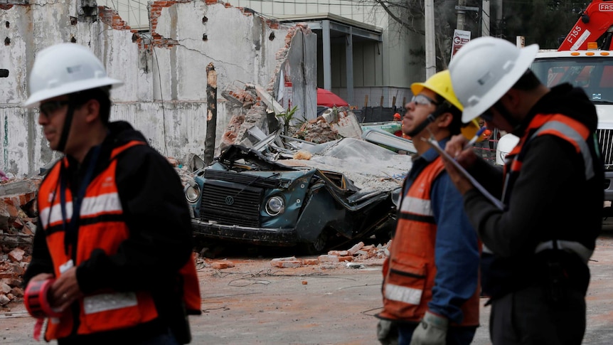 Three men in high-vis vests, slightly out of focus, stand in front of a damaged building and a crushed car.