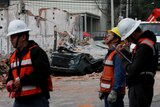 Three men in high-vis vests, slightly out of focus, stand in front of a damaged building and a crushed car.