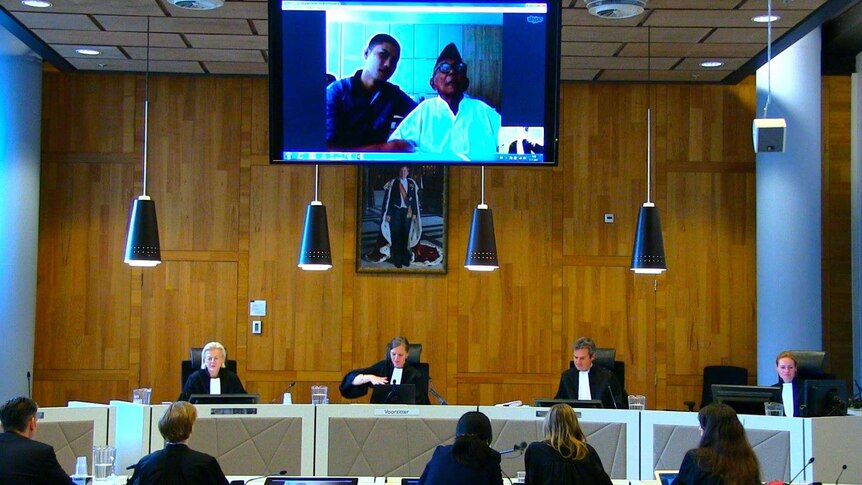 Four judges sit at a court bench, with a young man and an elderly gentleman appearing on a video screen above the desk.
