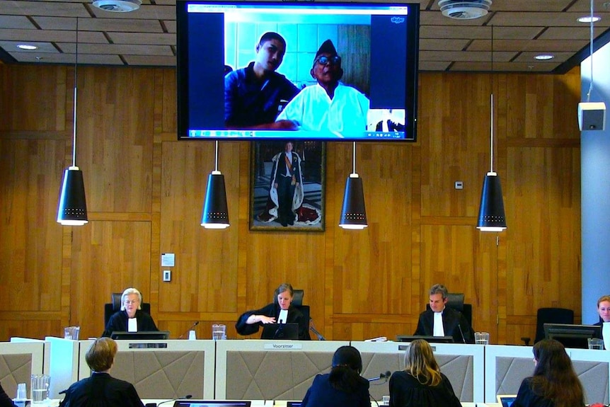 Four judges sit at a court bench, with a young man and an elderly gentleman appearing on a video screen above the desk.