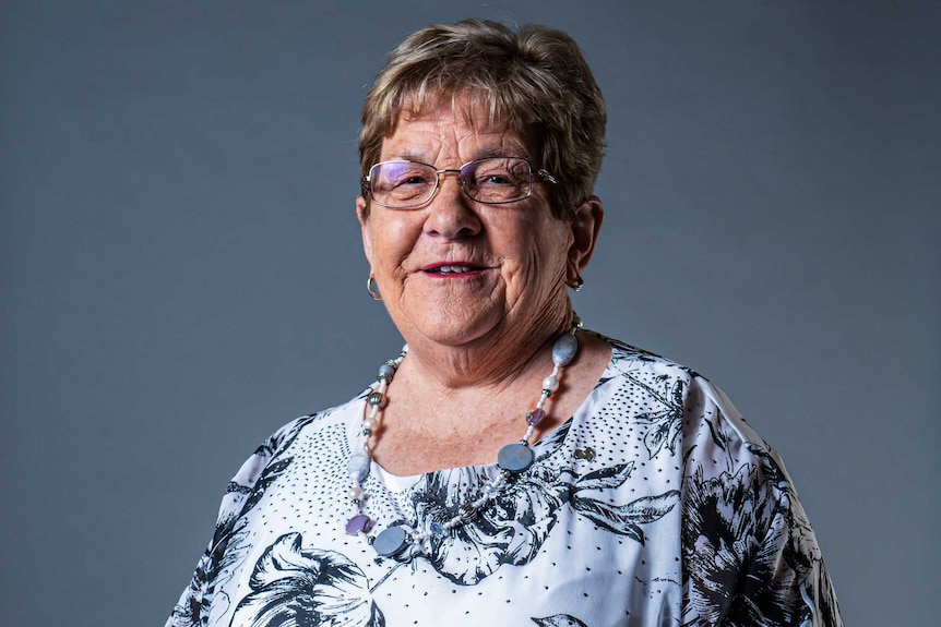 An elderly woman in a grey and white dress with matching necklace smiles at the canmera